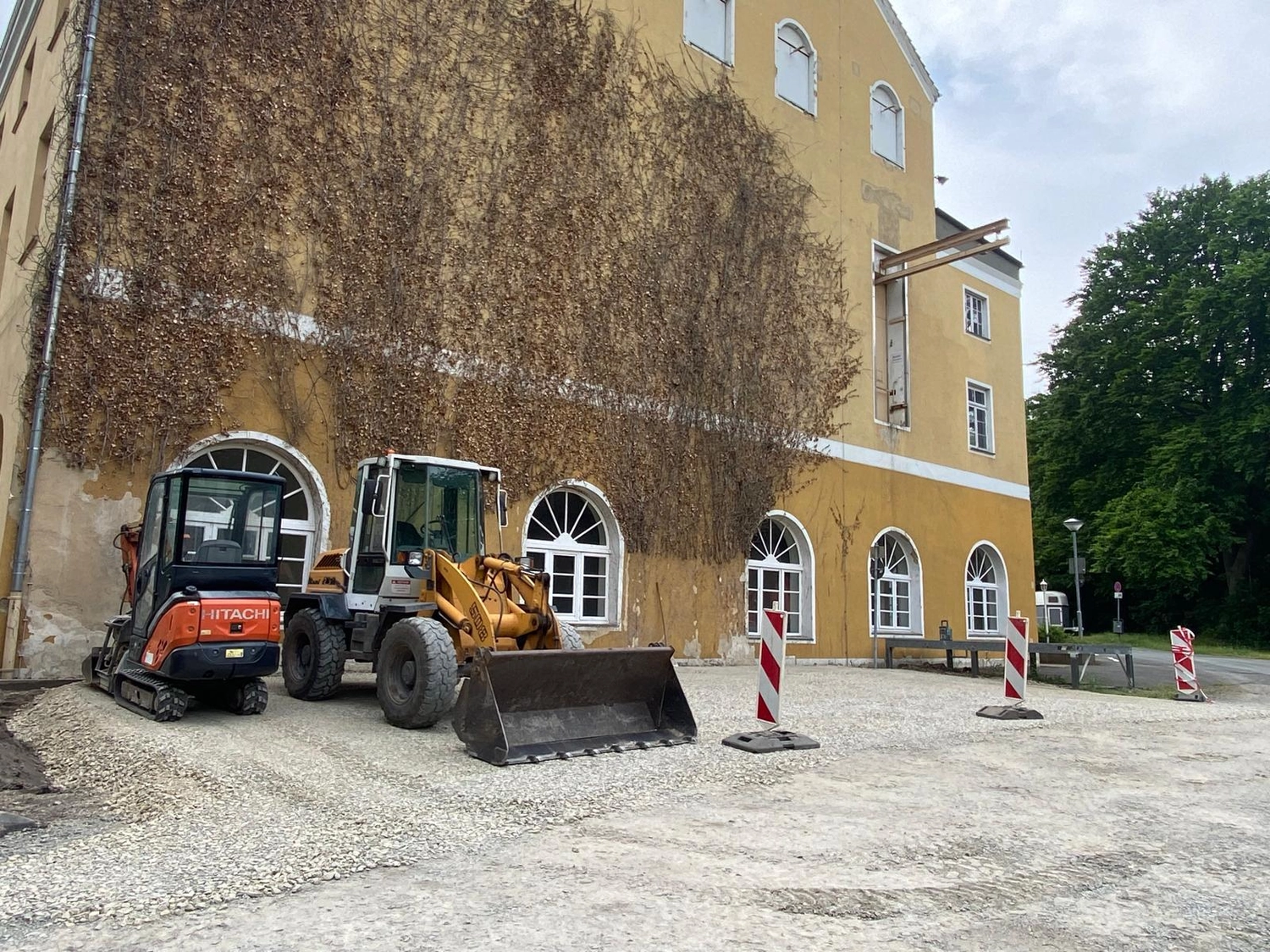 Stahlke - Tief und Landschaftsbau - Baumaschinen, darunter ein Hitachi-Bagger, parken vor einem gelben Gebäude.